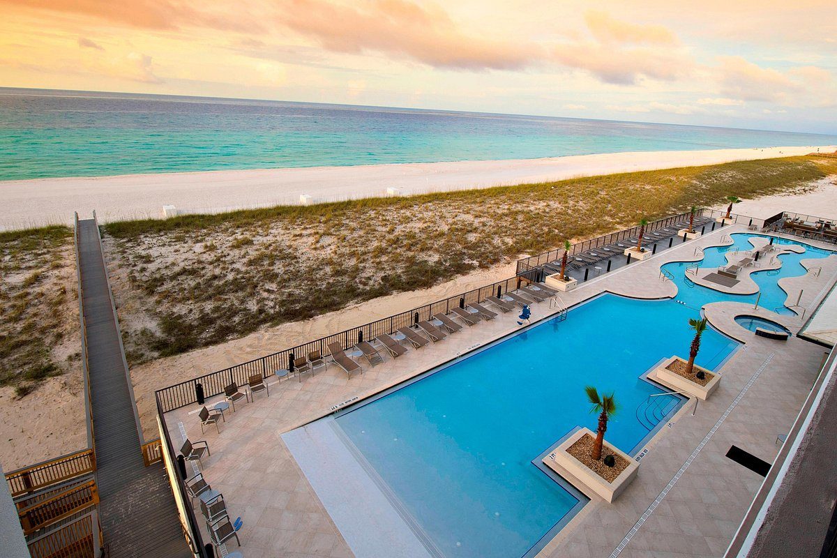 Pool bordered by palm trees, overlooking a sandy beach and calm ocean under a cloudy, colorful sky. Lounge chairs line the pool deck.