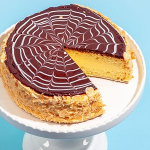 Cake sits on a white stand, with a slice removed, displaying yellow layers. Chocolate topping has white swirls; sides coated with almond slices.