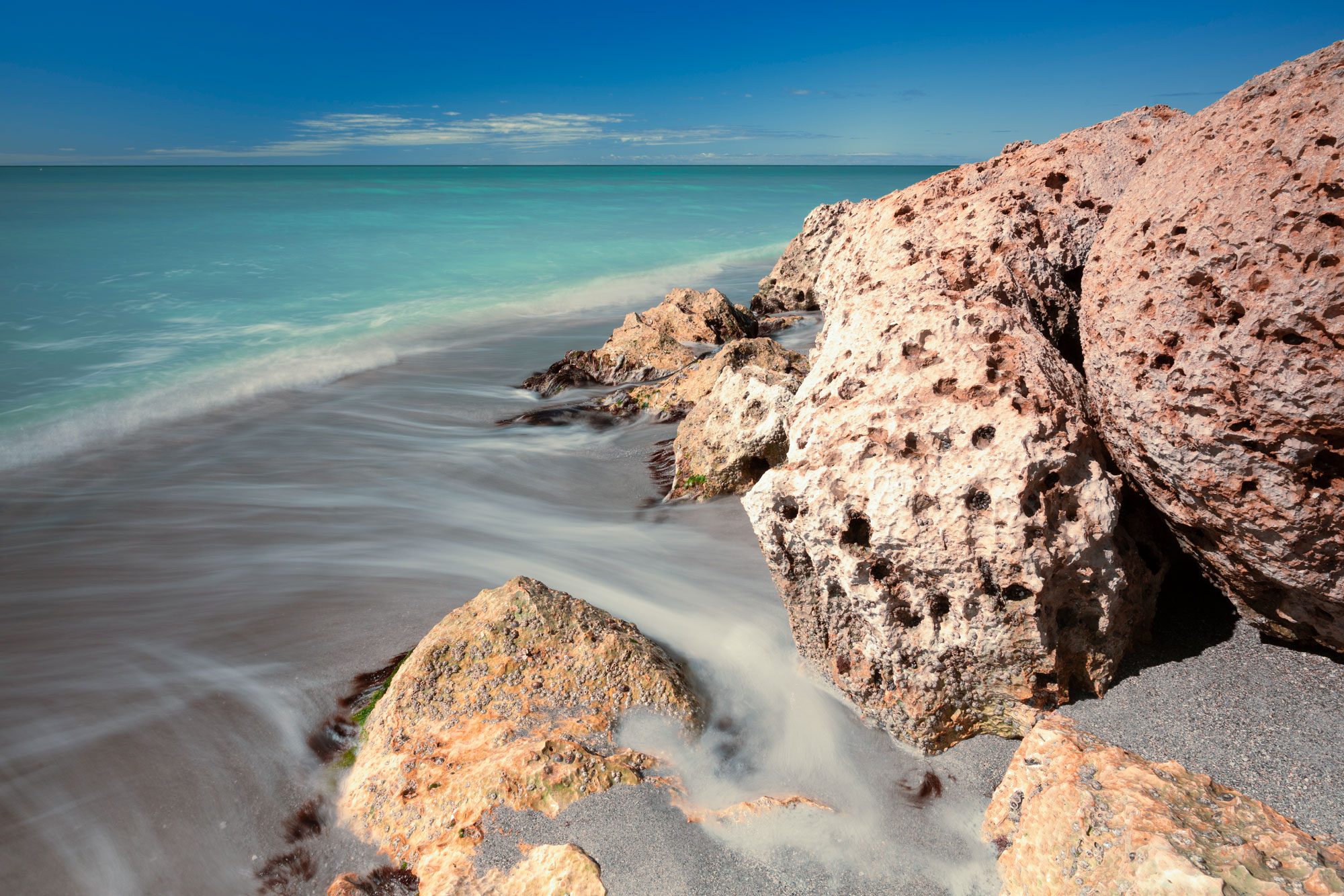 Waves wash over rugged rocks, creating a misty effect on a sandy beach under a clear blue sky.