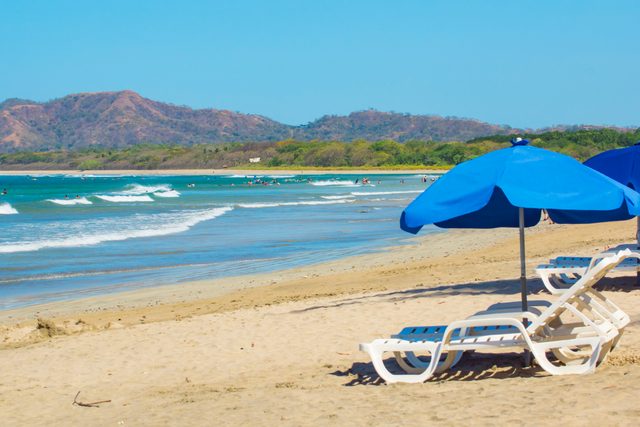Blue umbrellas shade beach chairs on sandy shore; waves gently roll in, framed by swimmers in ocean and distant green hills.