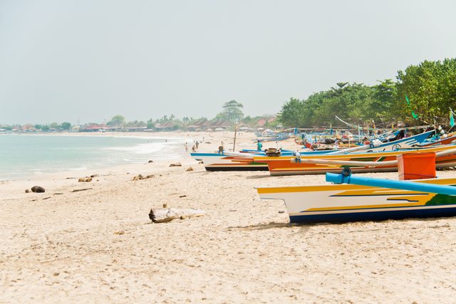 Colorful boats rest on sandy beach with scattered driftwood, ocean waves in the background, and green trees bordering a distant village.