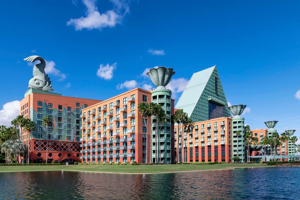 Building features pastel-colored towers with ornate roof sculptures, surrounded by palm trees and bordered by a calm lake, under a clear blue sky.