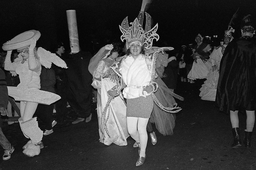 Person wearing elaborate headdress walks joyfully among others in costumes during a nighttime parade.