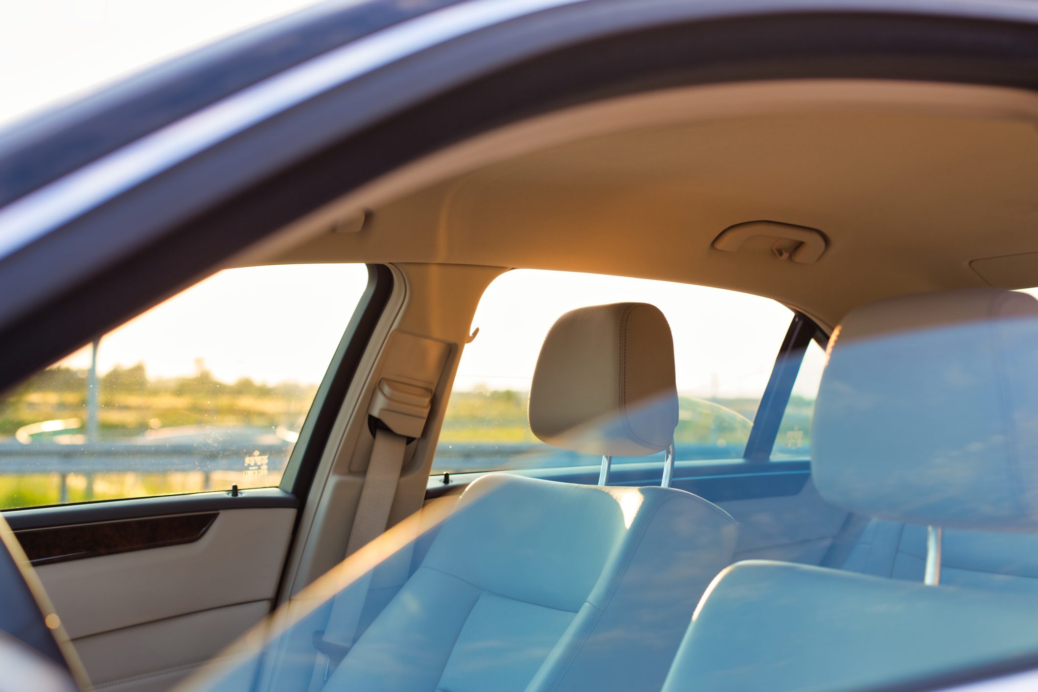 Car seats reflect sunlight inside a parked vehicle, with windows revealing a blurred outdoor view of greenery and a railing.
