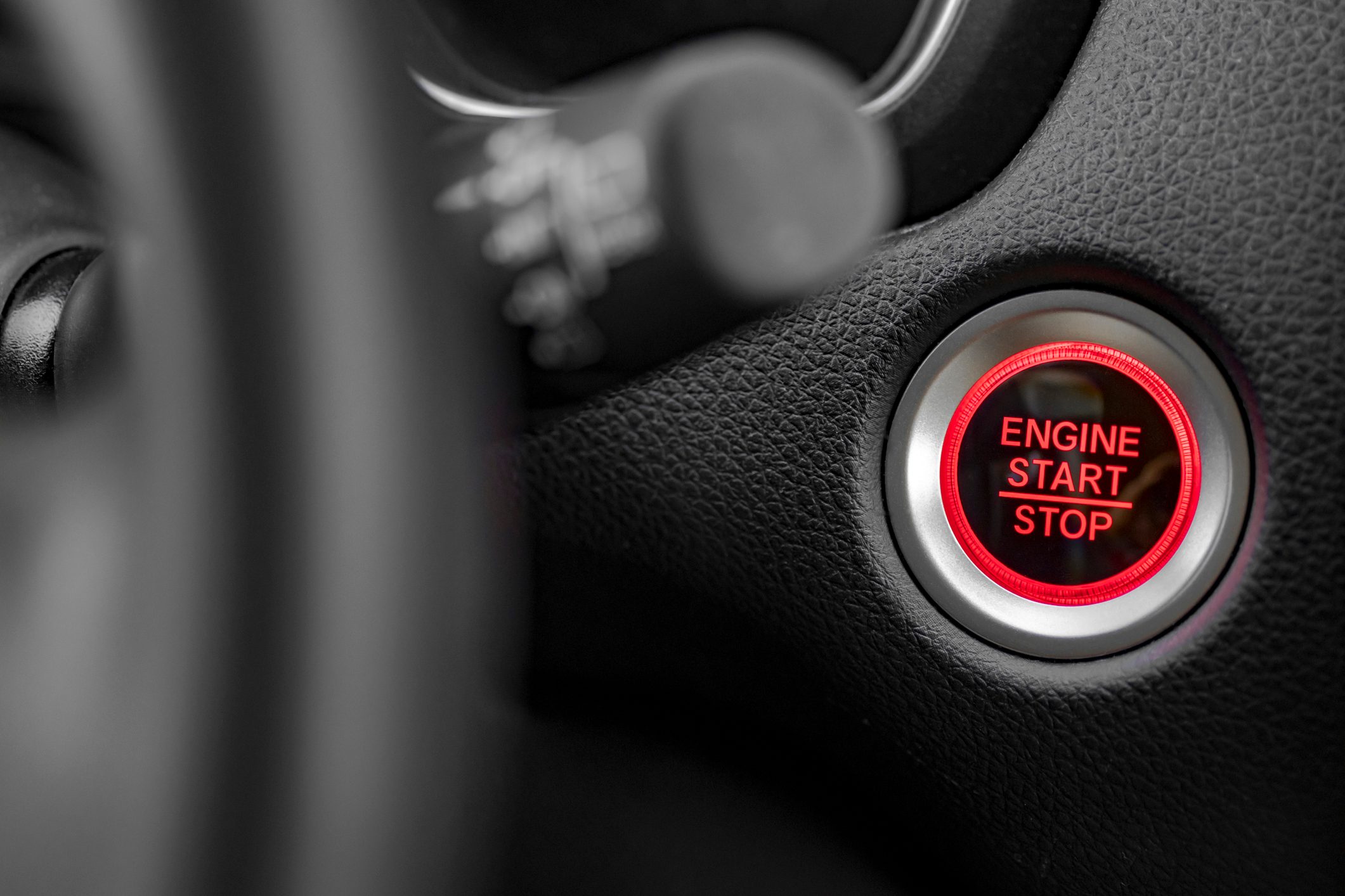 Button labeled "ENGINE START STOP" is illuminated, set within textured black car dashboard, next to steering wheel components.