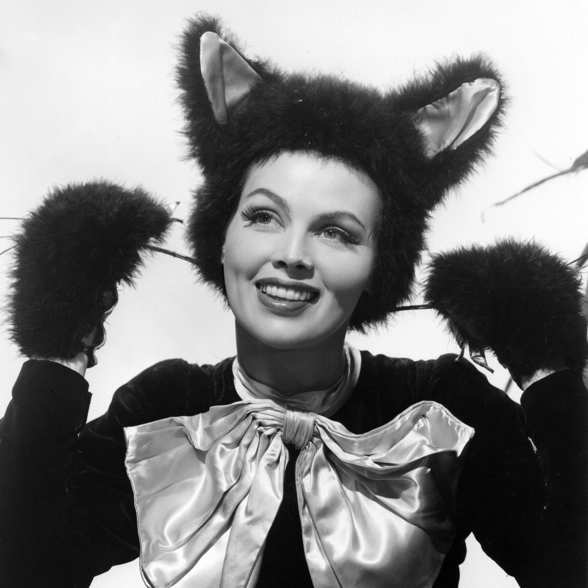 A person smiles wearing cat ears and paws, dressed in a large bow. They are in a photo studio with a plain background.