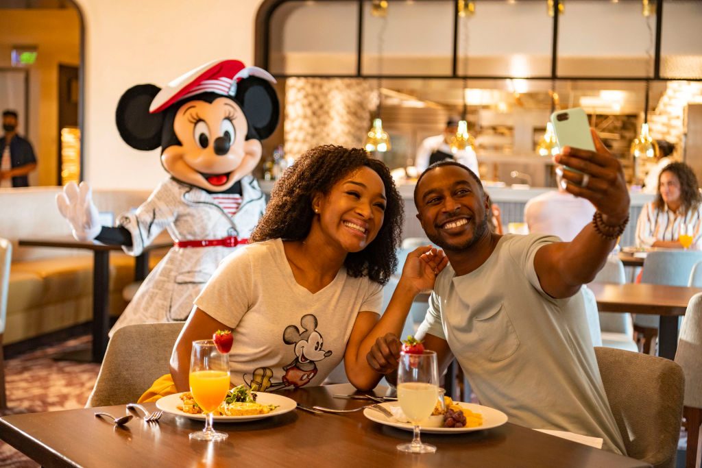 A couple takes a selfie in a restaurant, with Minnie Mouse posing behind them. The table has plates of food and drinks.