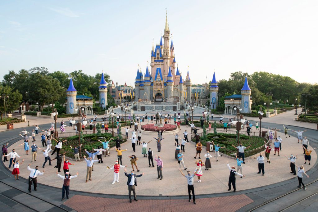 People standing with arms raised in front of a castle with blue turrets in a theme park, surrounded by gardens and paths.