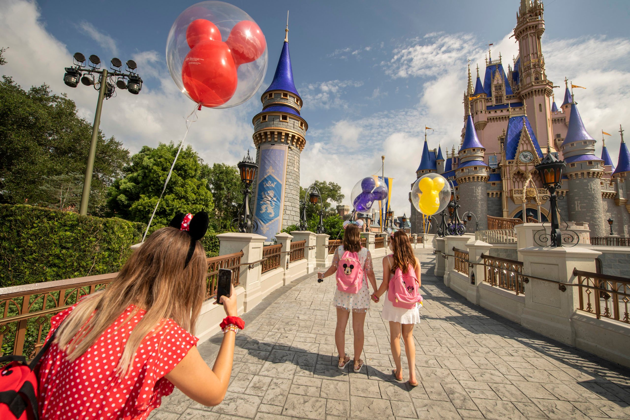 Balloons float and people walk towards a castle, with a woman capturing the moment with her phone on a sunny day.