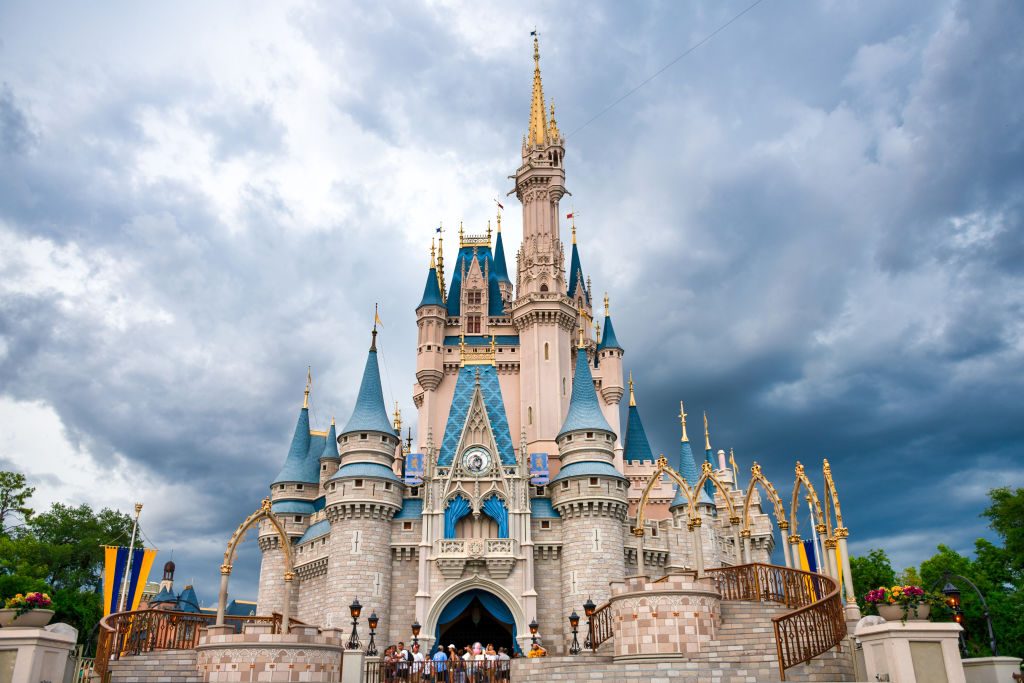A castle stands majestically, surrounded by decorative arches, under a dramatic cloudy sky, with tourists gathered at its base.