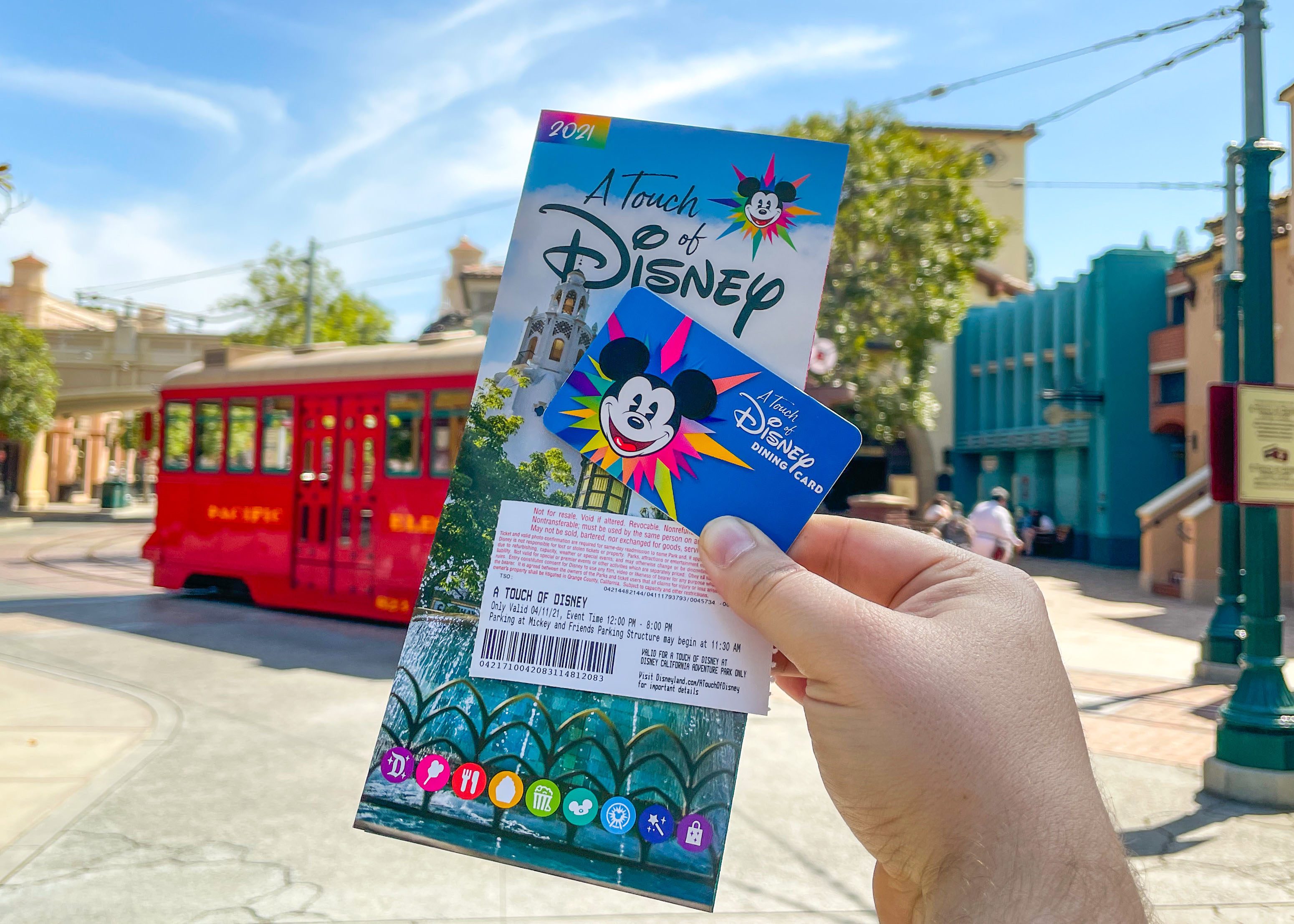 Hand holds Disney pass and dining card near red trolley on sunny street, surrounded by trees and buildings. Streetscape adds vibrant backdrop.