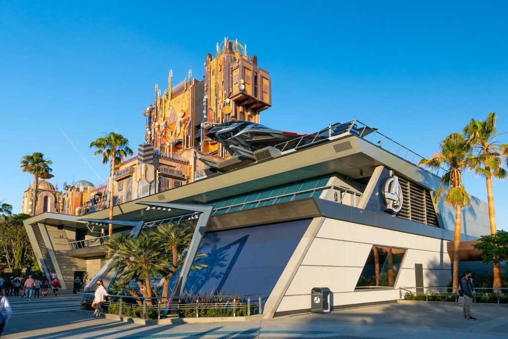 Futuristic building displays large Avengers logo, surrounded by palm trees and people walking, under clear blue skies.