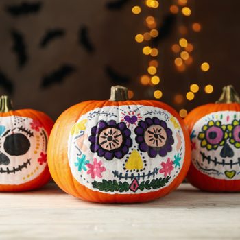 Pumpkins display colorful, intricately painted sugar skull faces; background features soft lights and silhouettes of bats on a dark wall.