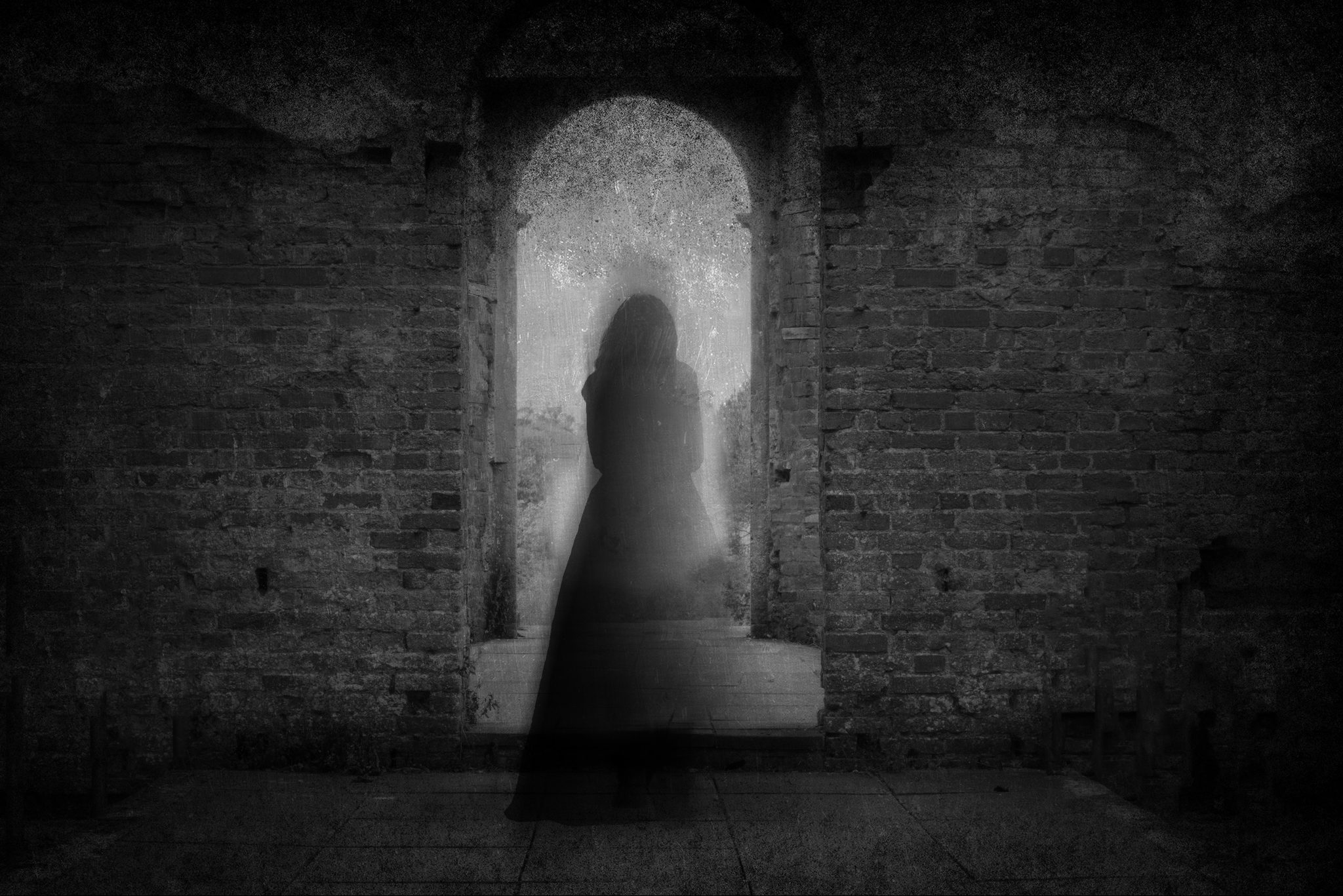 A spooky ghost of a woman in a dress, back to camera, framed by the archway of an old building. With a grunge, vintage, blurred edit.