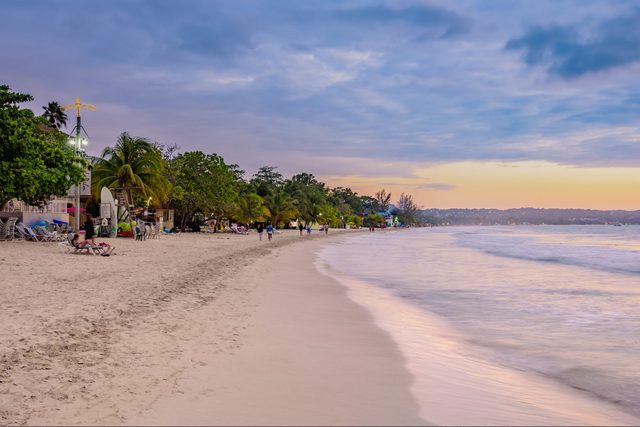 Beach at sunset, waves gently reaching shore, people strolling along sandy coastline lined with palm trees and lounge chairs, under a partly cloudy sky.