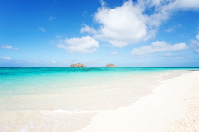 Beach stretches under clear blue sky, with turquoise waves gently lapping the sandy shore. Two small islands are visible in the distant horizon.