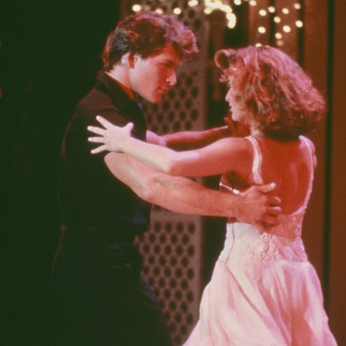 Two people dance closely, gazing at each other, in a warmly lit room with decorative lights in the background.