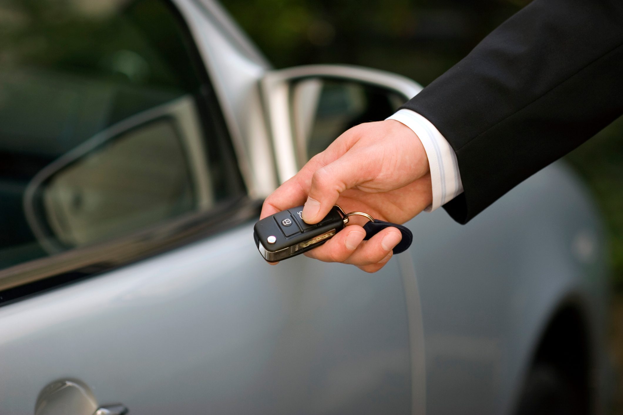 A hand holds a car key fob near a silver car in a parking context.