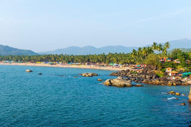Beachline populated with palm trees and small colorful huts, bordered by the sea, under a clear blue sky with distant hills in the background.