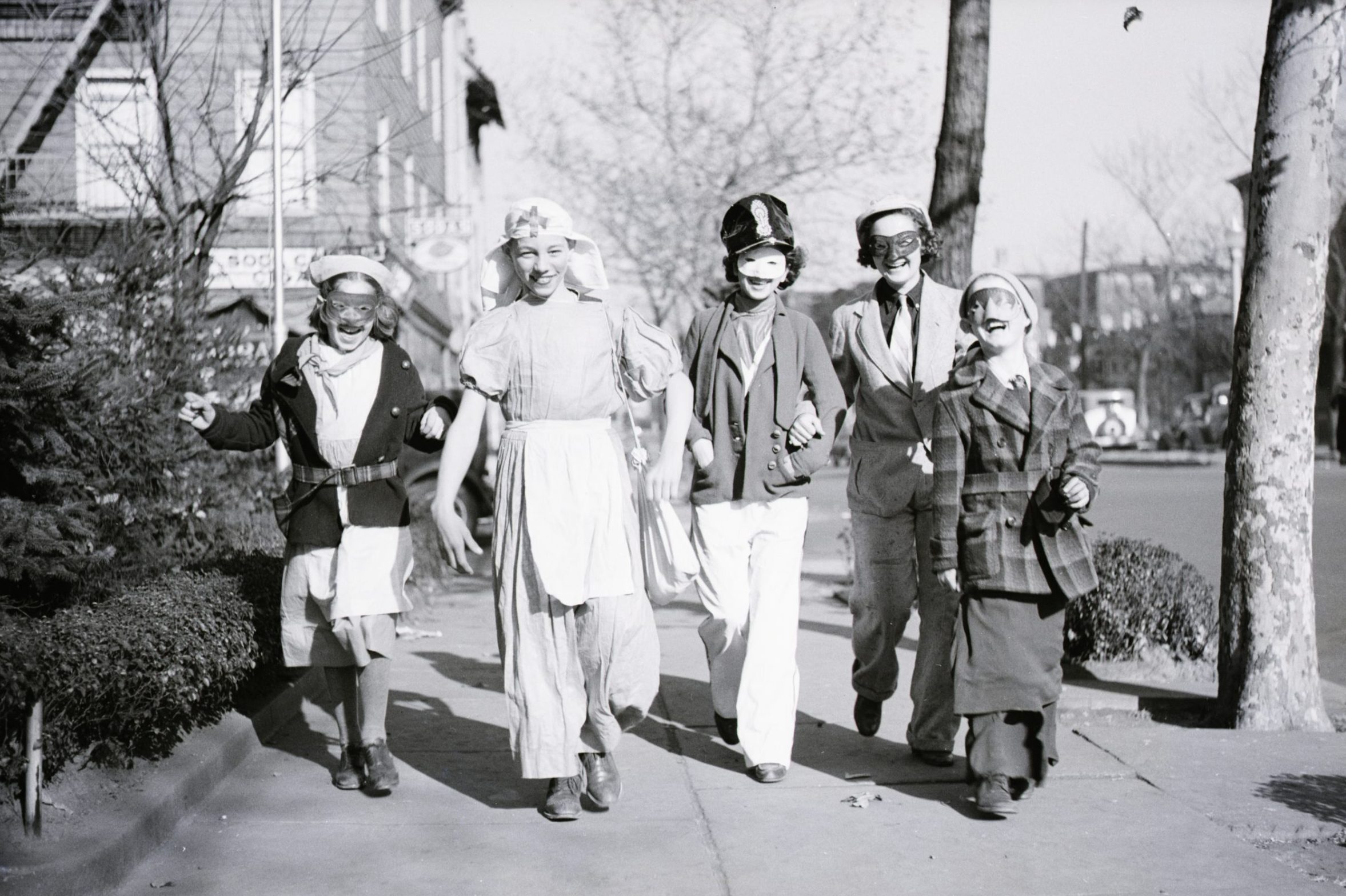 Five children walk together in costumes and masks, joyfully moving along a tree-lined urban sidewalk. Sparse trees and buildings outline the street background.