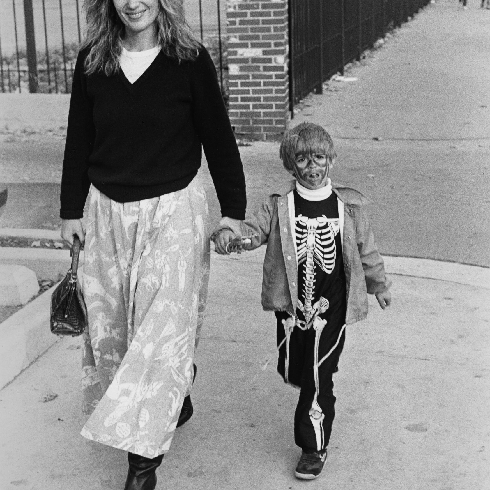 A child dressed as a skeleton walks hand-in-hand with an adult on a sidewalk next to a brick wall.