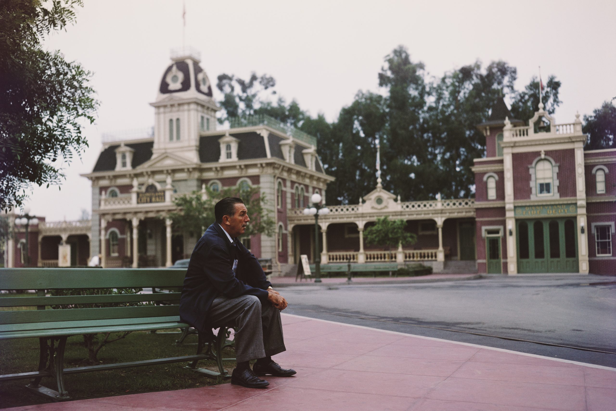 Man sits thoughtfully on a green bench; background features a quaint, ornate building with trees, suggesting a peaceful, picturesque setting.
