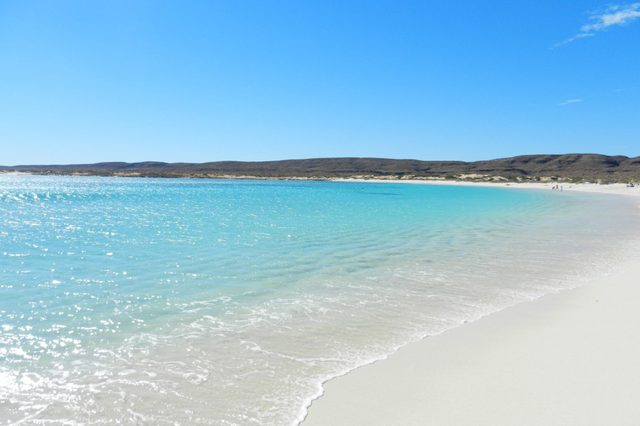 Turquoise waves gently reach a pristine, white sandy beach, set against a backdrop of distant hills under a clear blue sky.