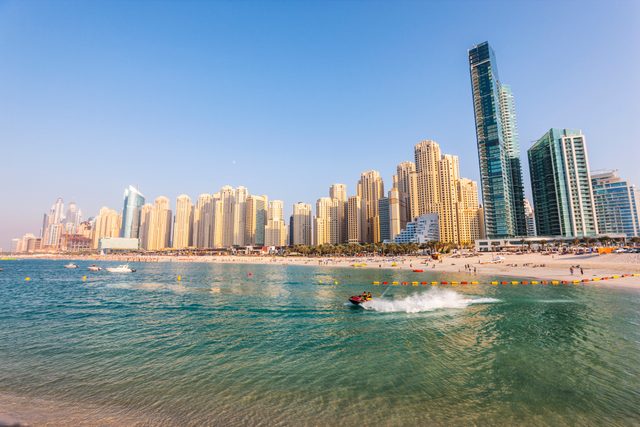 Jet ski speeds across turquoise water; sandy beach crowded with people; modern skyscrapers rise in the background under a clear blue sky.