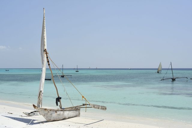 Sailboat rests on white sandy beach, facing calm turquoise waters, with other sailboats in the distant sea under a clear blue sky.