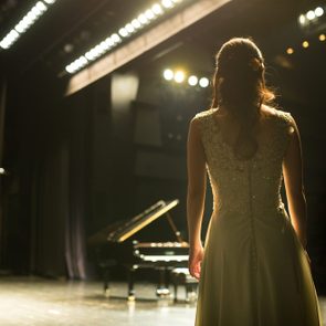 A woman stands, back to camera, facing a grand piano on a dimly lit stage.