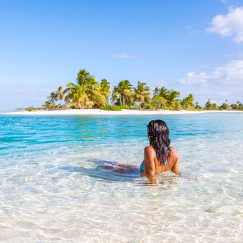 Person relaxes in shallow turquoise water, facing a tropical island with palm trees under a clear blue sky.