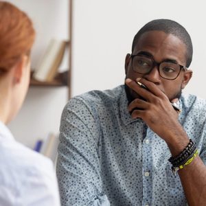 Man thinking intently, hand on face, sitting across from someone, in an office setting with a bookshelf in the background.