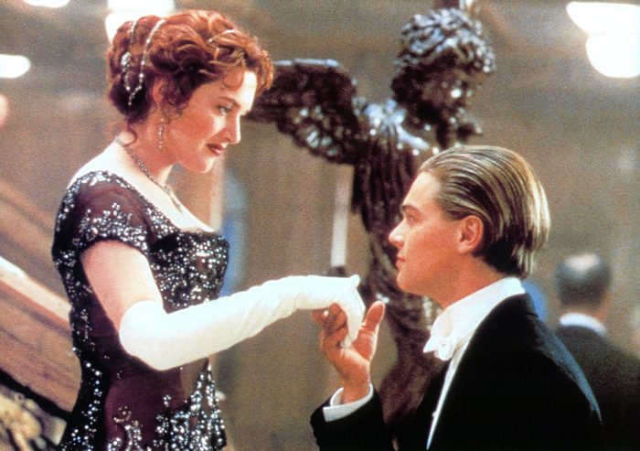 A woman in a beaded dress and gloves offers her hand to a man in a tuxedo, set indoors with a decorative sculpture.