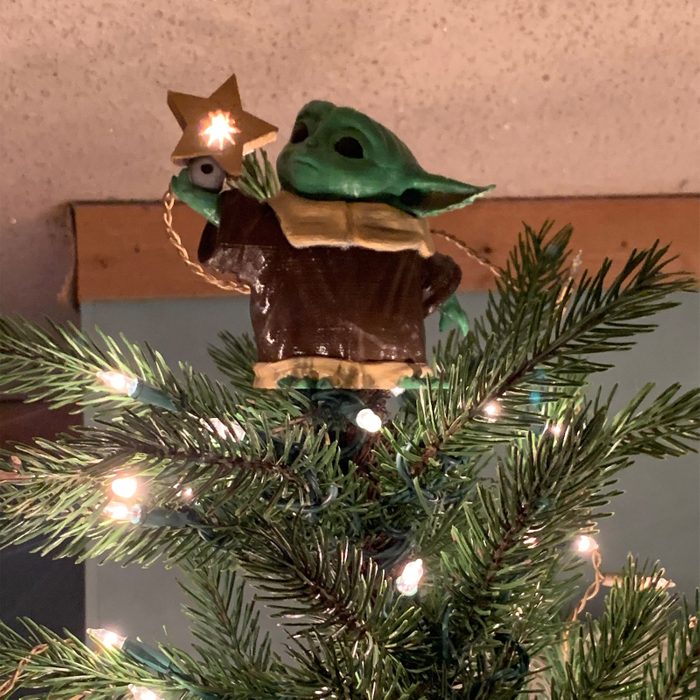 Figure tops tree, holding a glowing star, surrounded by green branches decorated with white lights.