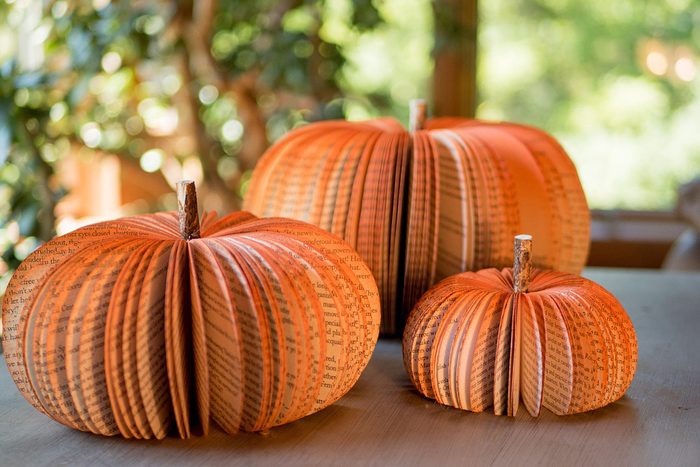 Three book page pumpkins sit on a table in a sunlit room, surrounded by greenery outside the window.