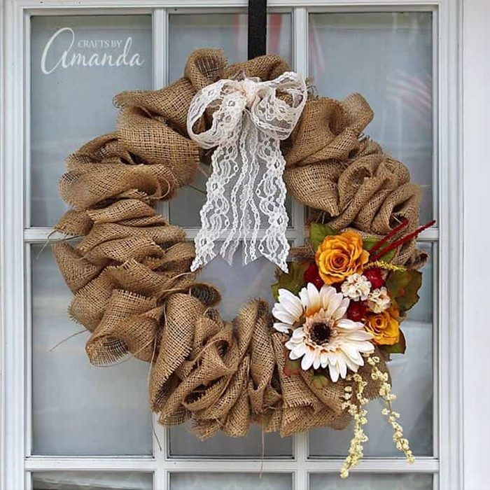 Burlap wreath hangs on a window, adorned with a lace bow and clustered flowers, including sunflowers and roses in warm colors. Text: 
