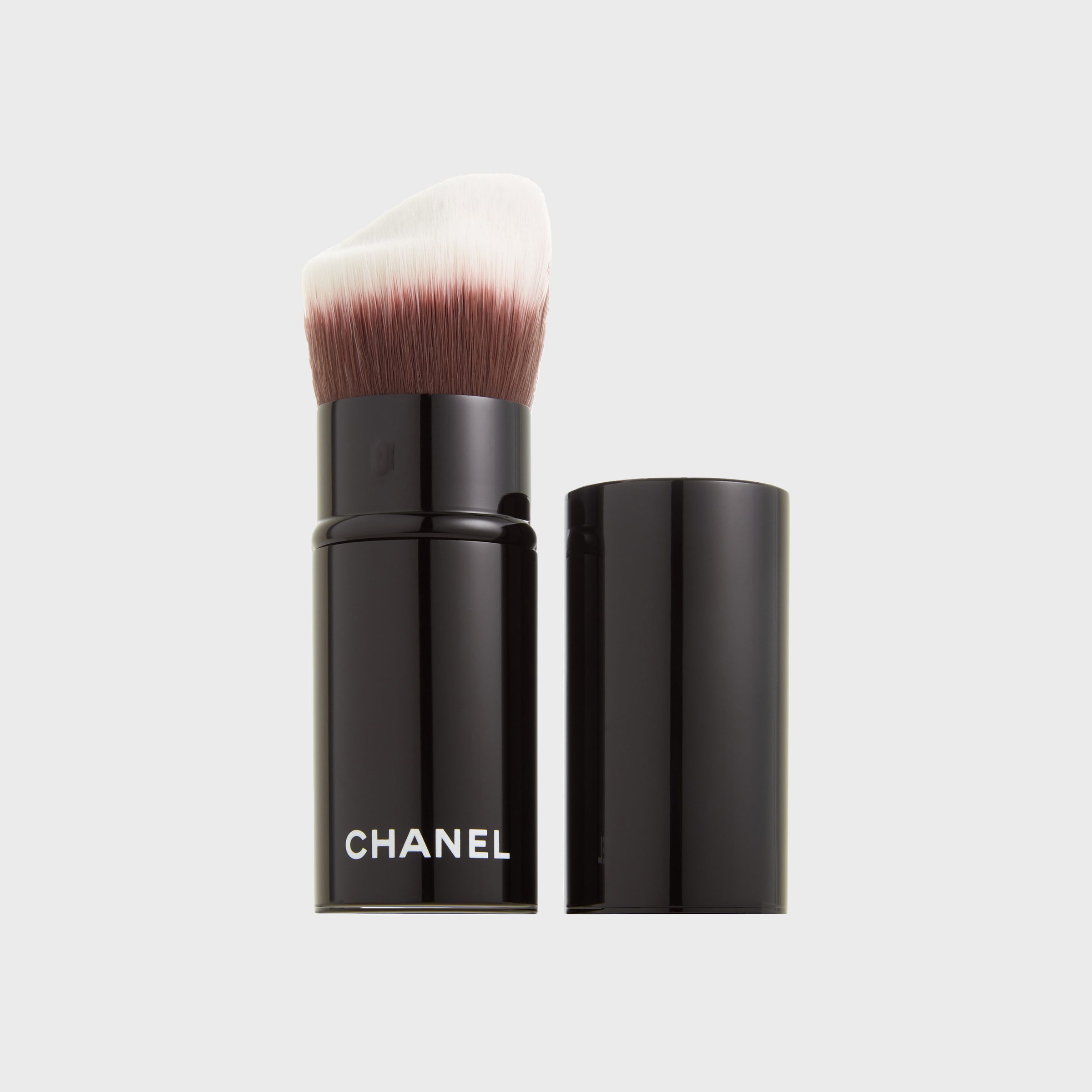 Makeup brush stands upright with bristles exposed, next to its black cap. "CHANEL" is printed on the brush handle. White background.