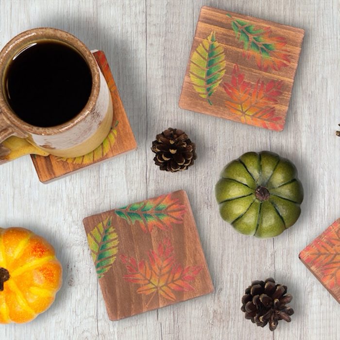 Coasters with colorful leaf designs sit on a wooden table alongside pine cones, a cup of coffee, and small decorative pumpkins.