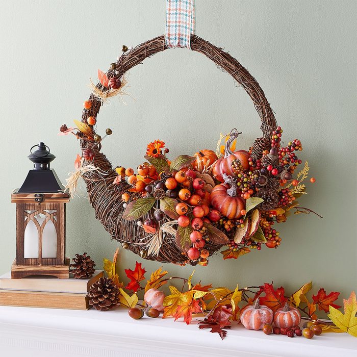 Wreath adorned with pumpkins and berries hangs against a green wall, accompanied by a wooden lantern and autumn leaves on a mantel.