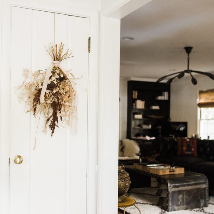 A dried floral arrangement hangs on a white door; beyond, a living room features a dark bookshelf, couch, and a metal coffee table.