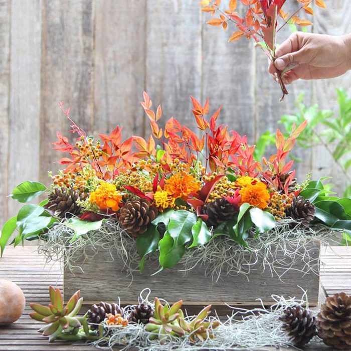 Floral centerpiece, decorated with autumn leaves and pinecones, being arranged in a rustic wooden box on a wooden table outdoors.