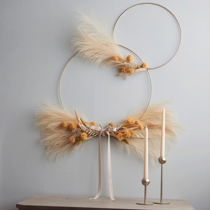 Decorative hoops hold dried pampas grass and yellow flowers, accented with ribbons, above two tall, slender candles on a wooden surface against a plain wall.