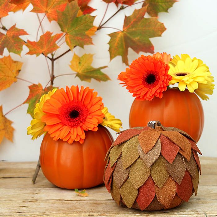 Pumpkins display bright orange and yellow flowers on wood surface, surrounded by autumn leaves on branches in a festive fall arrangement.