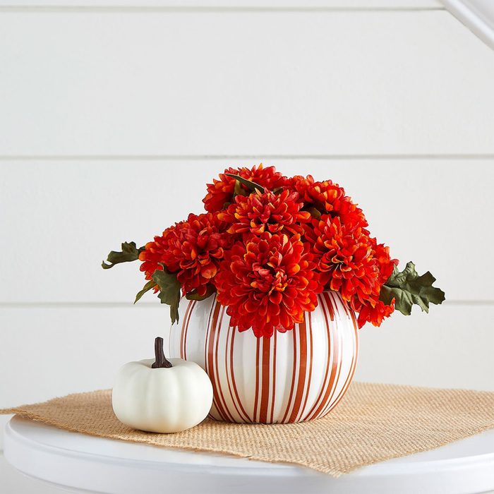 Striped vase holds red flowers, sitting beside a small white pumpkin on a round table covered with burlap, against a white shiplap background.