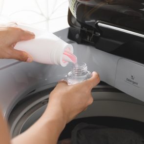 Bottle pours pink liquid into measuring cap beside washing machine compartments labeled 