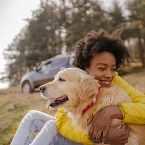 Person hugging a golden retriever, smiling in a grassy field with trees and a parked car in the background.