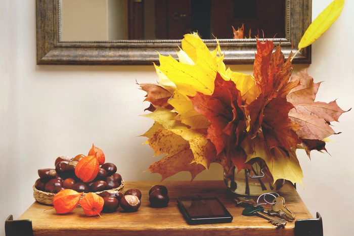 Autumn leaves arranged in a vase, chestnuts and orange husked fruits in a basket, on a wooden table below a framed mirror.