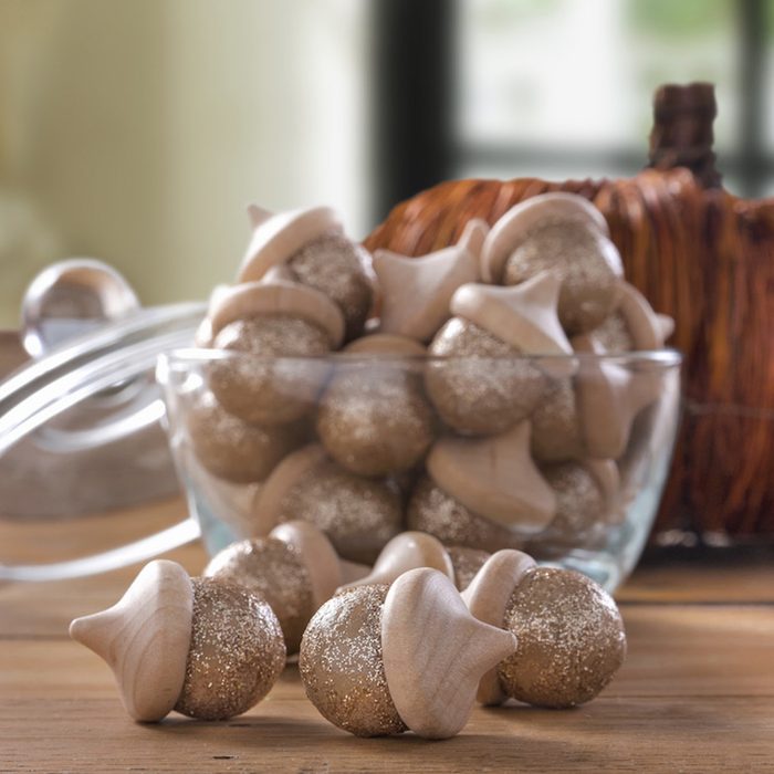 Glittery, wooden acorns sit inside a glass bowl on a wooden table, with a blurred background of a window and decorative pumpkin.