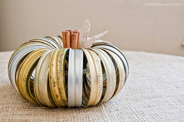 Jar lids form a pumpkin shape, with cinnamon sticks and burlap as the stem, resting on burlap fabric.