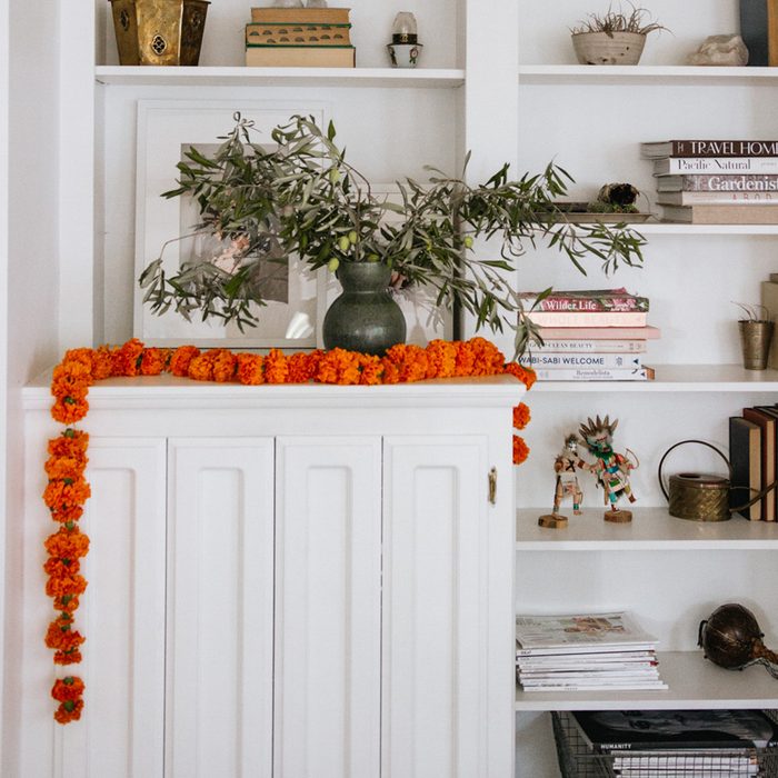 Green vase holds branches, sits on a white cabinet adorned with orange marigold garland; surrounded by books and decor on shelves.